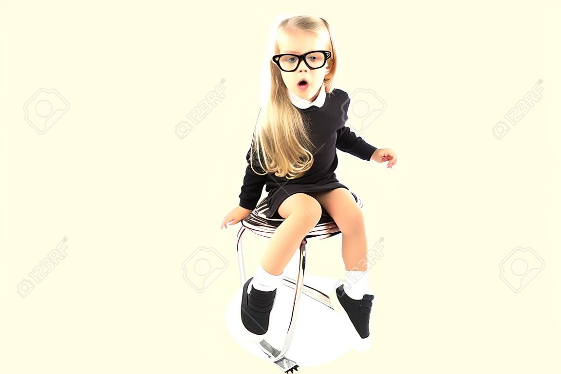 Isolated on white, pretty caucasian blonde little girl in black dress, glasses, black socks and white shoes sitting, look at camera, amazed