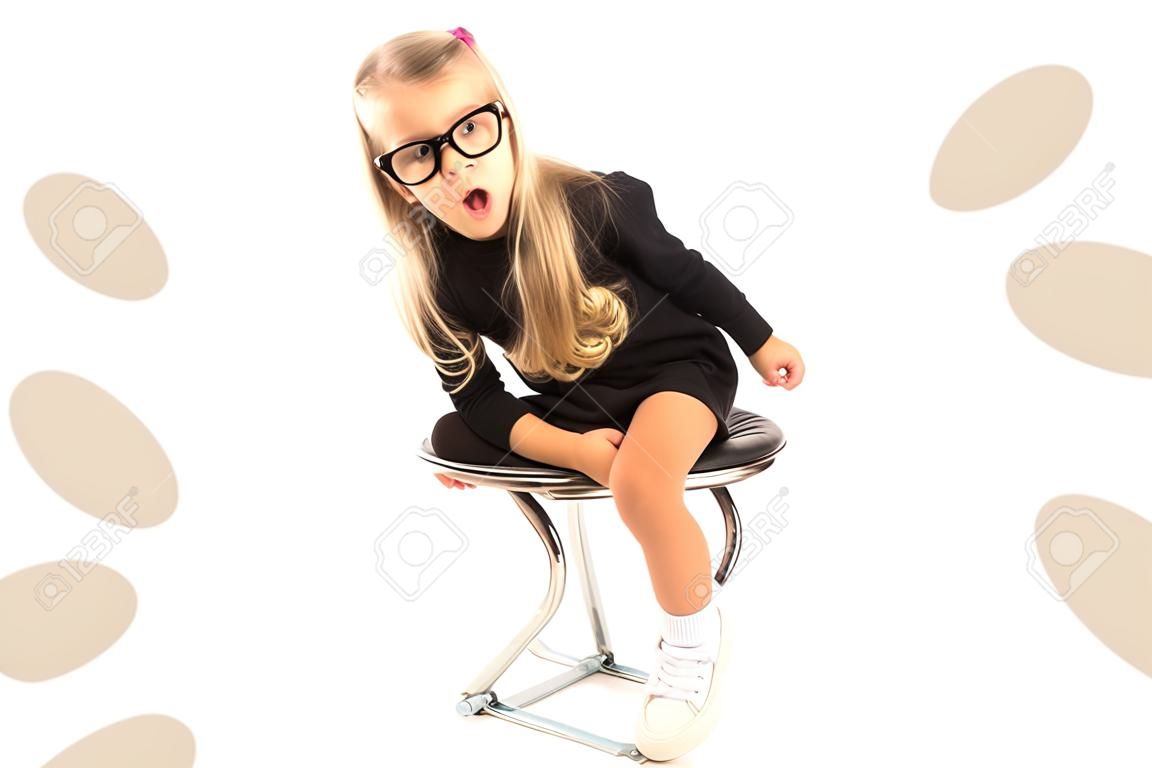 Isolated on white, pretty caucasian blonde little girl in black dress, glasses, black socks and white shoes sitting, look at camera, amazed