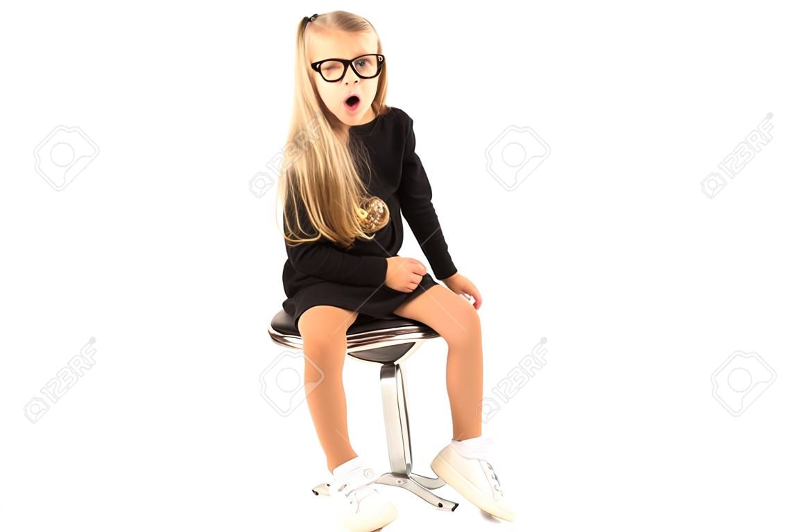 Lokalisiert auf Weiß, recht kaukasisches blondes kleines Mädchen im schwarzen Kleid, in den Gläsern, in den schwarzen Socken und in den weißen Schuhen, die sitzen, Blick in die Kamera, erstaunt
