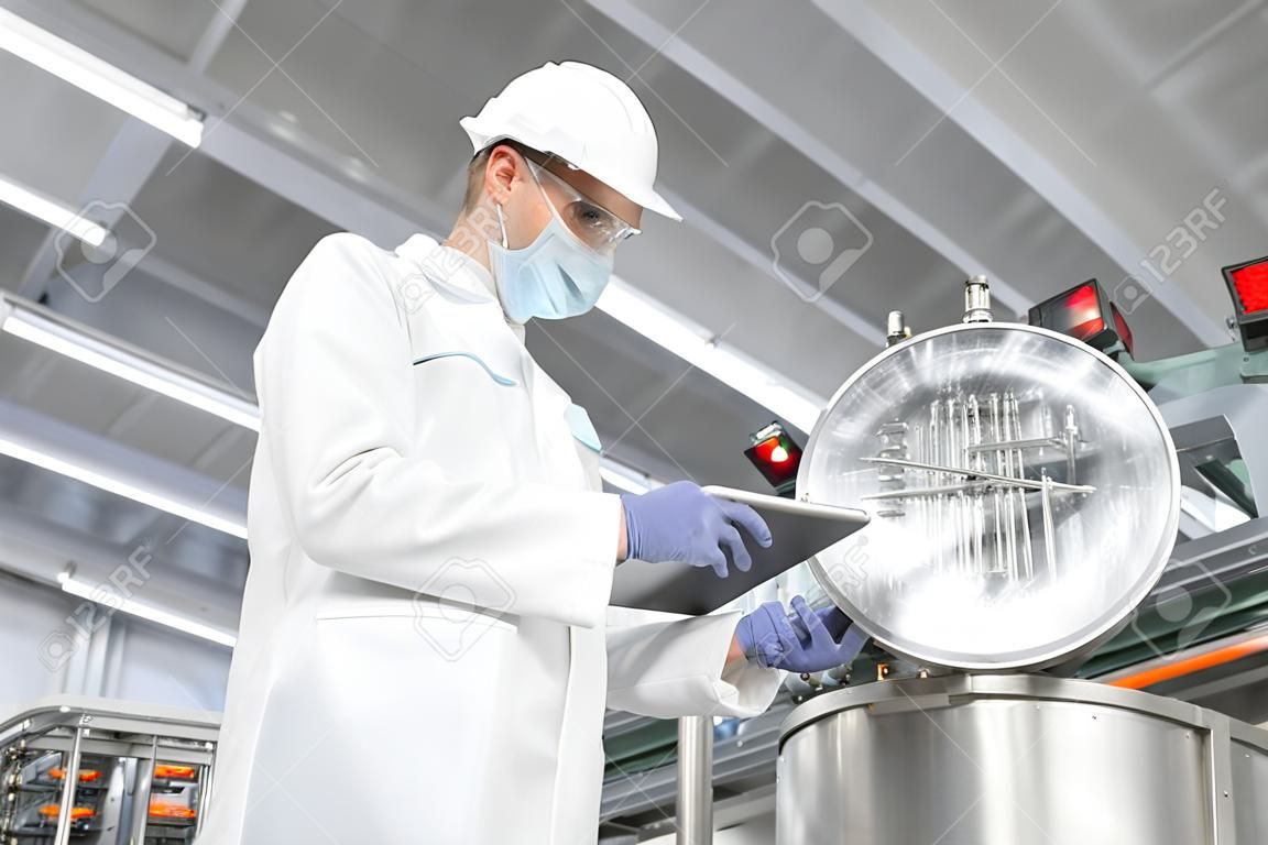 Technologist in a cap, mask and white coat is in the production shop and holding a digital tablet. inspector removes the indicators at the dairy plant. Engineer keeps statistics on production