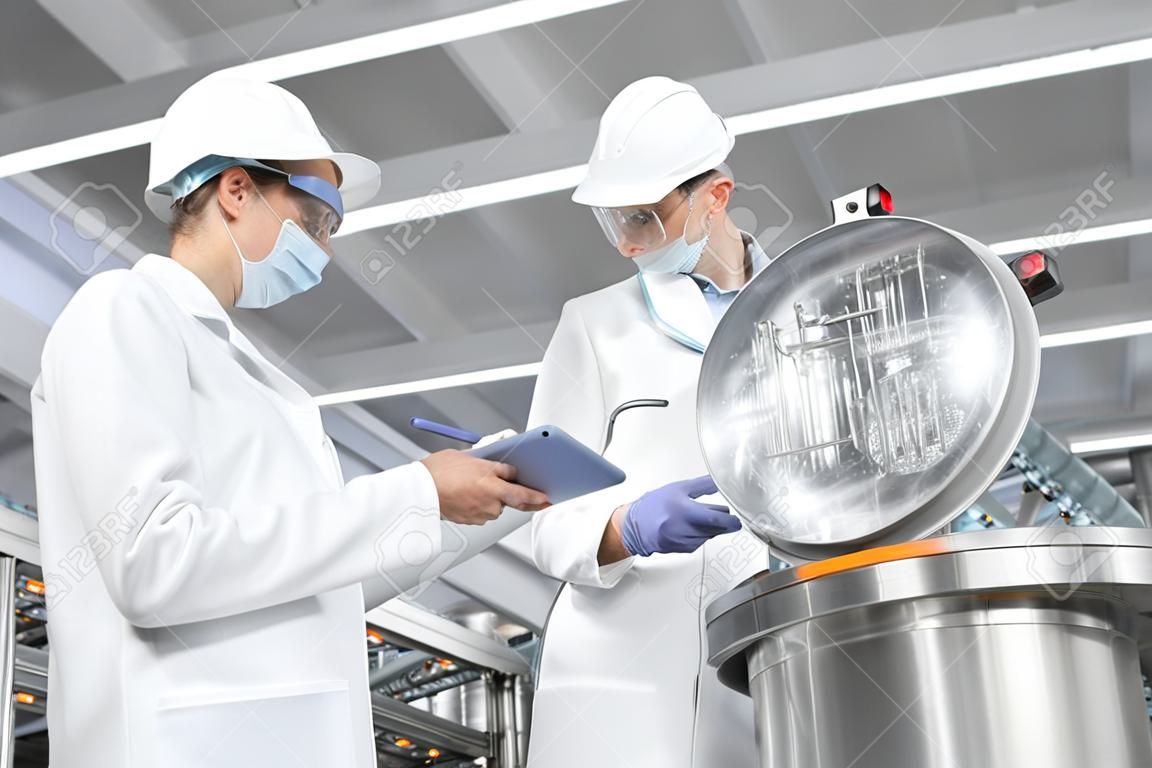 Technologist in a cap, mask and white coat is in the production shop and holding a digital tablet. inspector removes the indicators at the dairy plant. Engineer keeps statistics on production