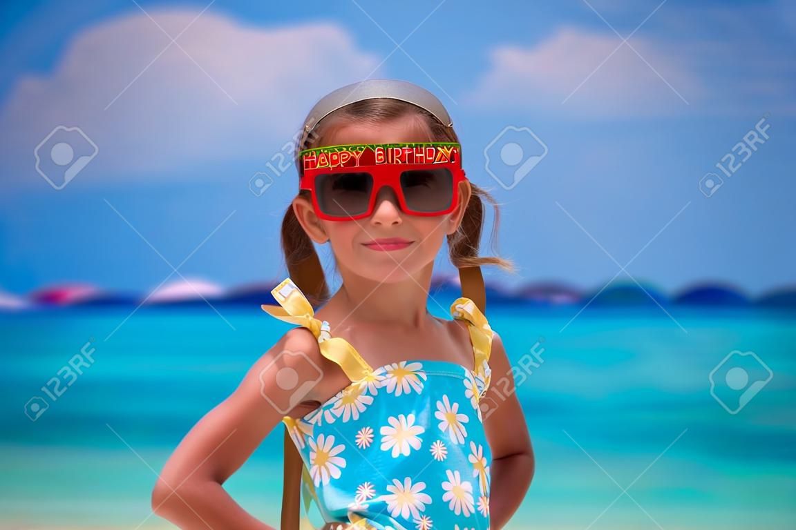 Adorable little girl in Happy Birthday glasses smiling and having fun at white beach