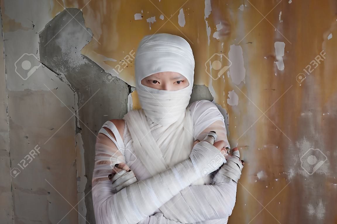 Halloween image, mummy in bandages, risen dead legendary character. young woman in the form of a mummy wrapped in bandages, against the textured old wall
