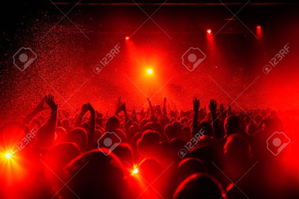 Uma sala de concertos lotada com luzes alaranjadas e amarelas do palco da cena, desempenho do show do rock, com silhueta das pessoas, explosão colorida do confete disparada no ar da pista de dança durante o festival de concerto