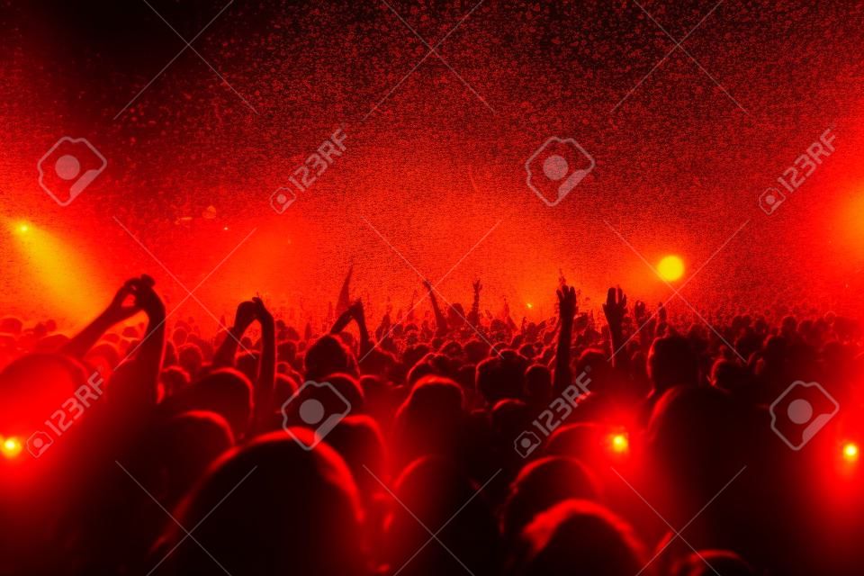 Uma sala de concertos lotada com luzes alaranjadas e amarelas do palco da cena, desempenho do show do rock, com silhueta das pessoas, explosão colorida do confete disparada no ar da pista de dança durante o festival de concerto