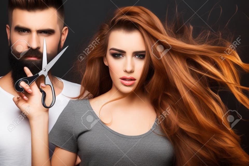 年轻漂亮的几个美丽时尚的女理发师拿着剪刀切割和英俊的男子在黑色背景上的水平图像工作室长胡须放大图