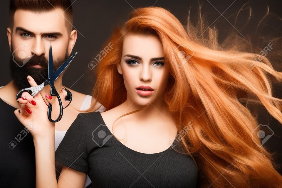 年轻漂亮的几个美丽时尚的女理发师拿着剪刀切割和英俊的男子在黑色背景上的水平图像工作室长胡须放大图