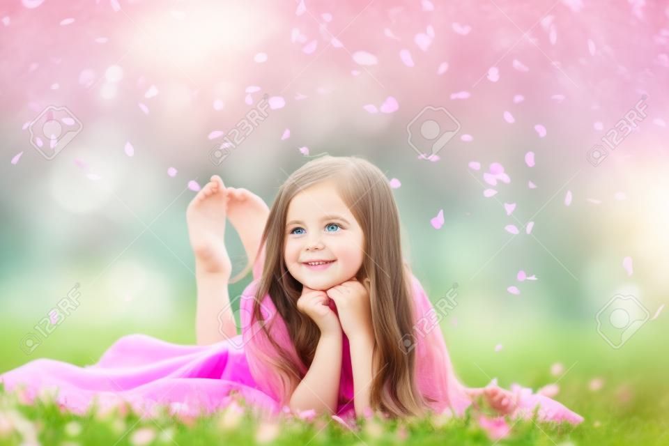 漂亮的小女孩在粉紅色的衣服，長黑髮頭髮和春季盛開的花瓣在綠色草地上微笑的臉躺在戶外赤腳