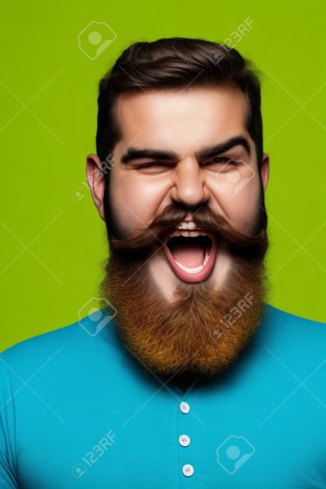 handsome bearded man with long lush beard and moustache on shouting face in yellow shirt in studio on blue background