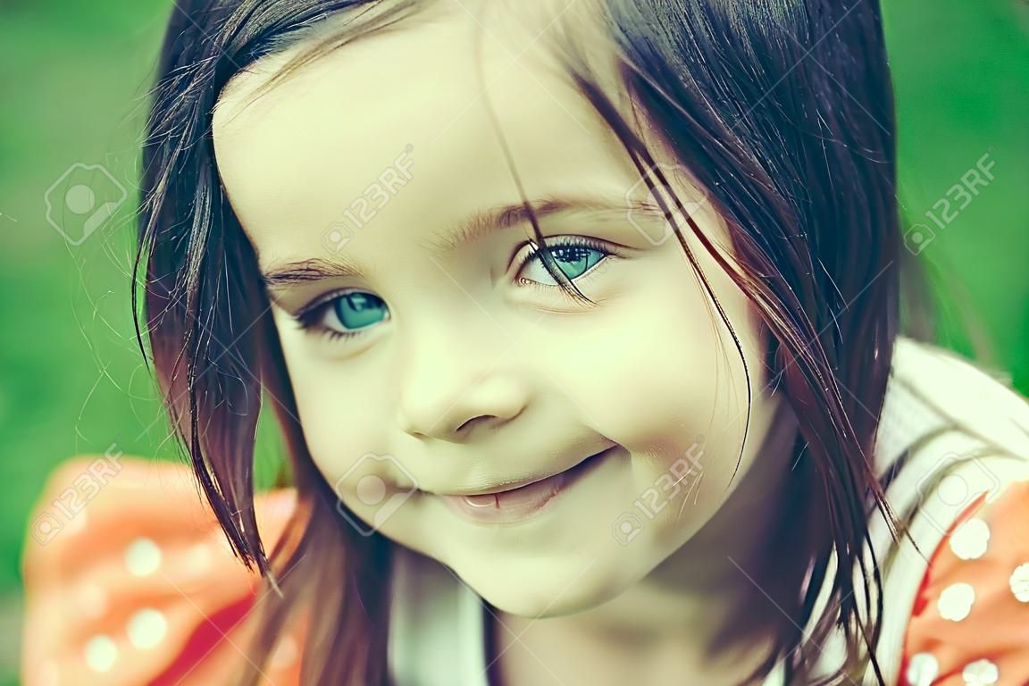 Smiling face of little cute happy girl child with blue eyes and brunette hair outdoor closeup on blurred green background