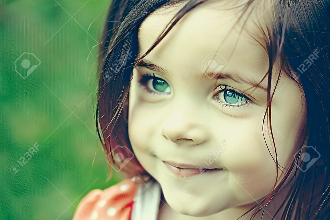 Улыбающееся лицо маленькой милой счастливым девочек с голубыми глазами и темные волосы на открытом воздухе крупным планом на размытом зеленом фоне