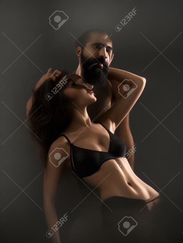 joven pareja de la mujer con la cara muy morena de pelo negro en sujetador erótico en el cuerpo sexy y guapo hombre de la barba con la barba larga en el estudio sobre fondo gris oscuro