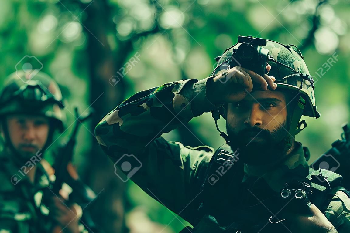 Young soldier man with beard on grime serious face in ammunition and helmet holding camera in his hands on background of soldiers in forest