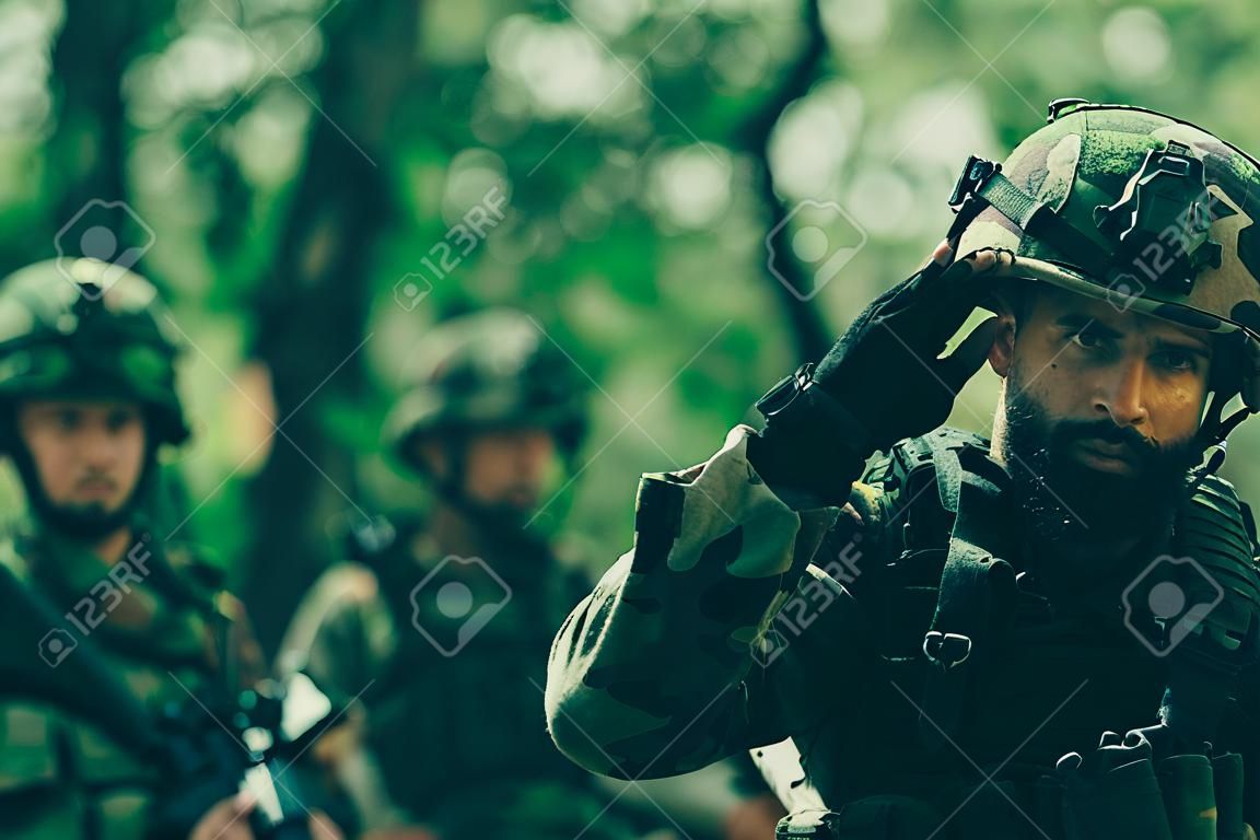 Young soldier man with beard on grime serious face in ammunition and helmet holding camera in his hands on background of soldiers in forest