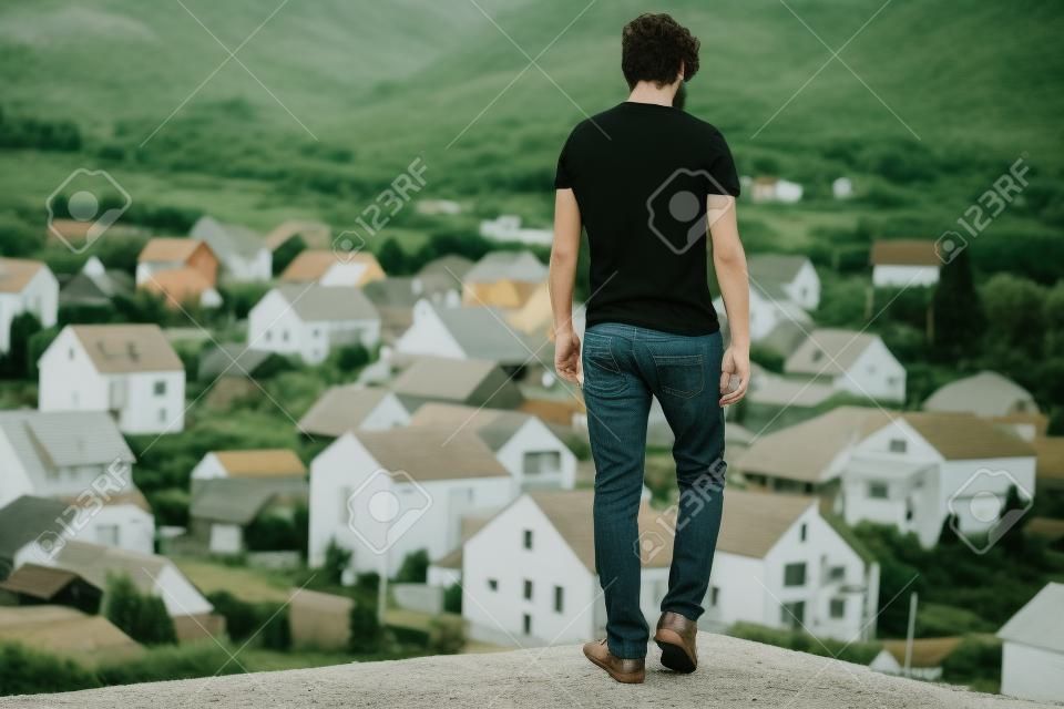 Young man brunette in black tshirt and jeans casual wear enjoys rural scenery from mountain