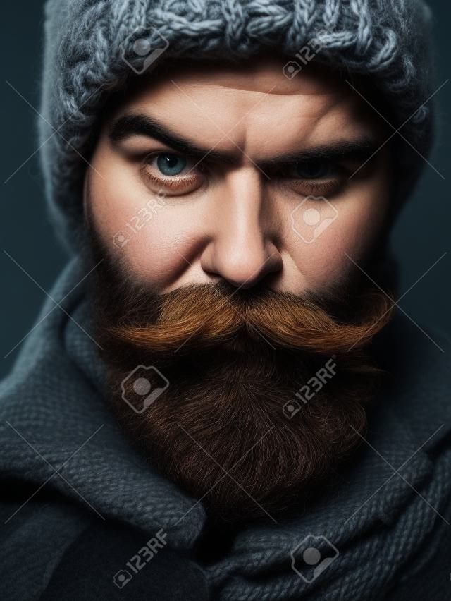 Fruncir el ceño barbudo hombre con barba y bigote con estilo hipster masculino en cálido sombrero de punto y abrigo al aire libre sobre fondo oscuro
