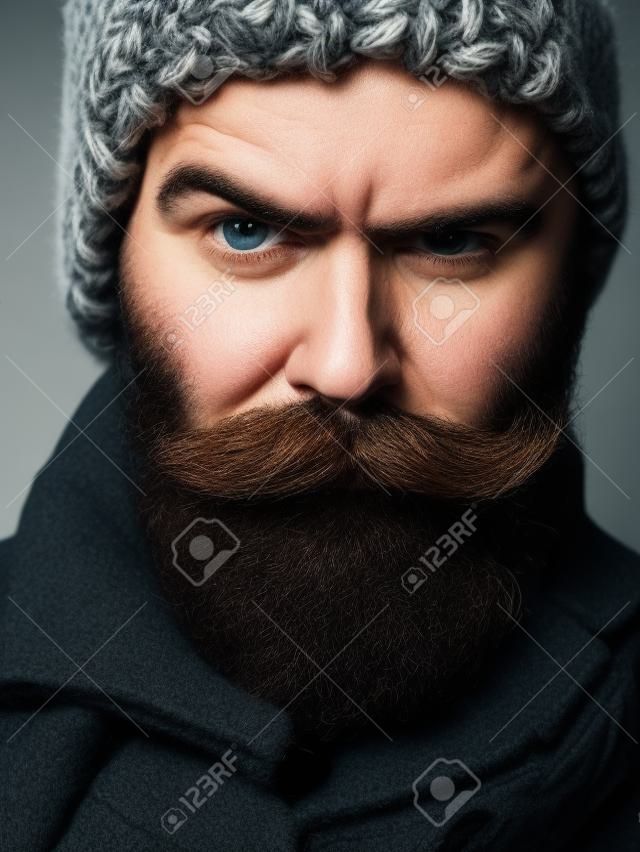 Stirnrunzeln bärtigen Mann mit Bart und Schnurrbart stylish hipster männlich in warmen gestrickten Hut und Mantel im Freien auf dunklem Hintergrund