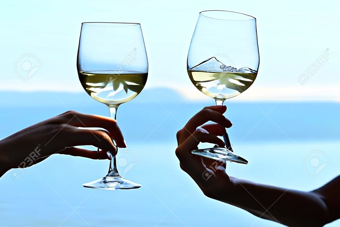 Hands of male and female clinck wineglasses of white wine on blurred seascape background.