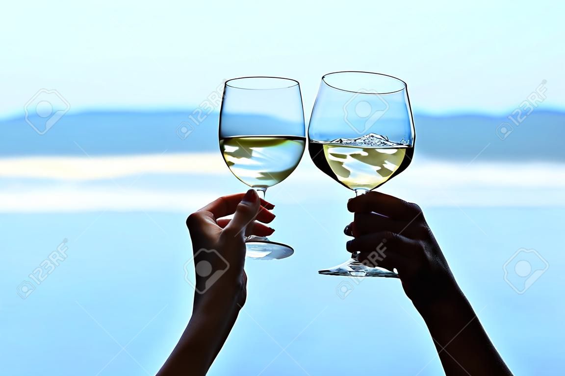 Mani dei bicchieri di vino del clinck del maschio e della femmina di vino bianco sul fondo vago di vista sul mare.