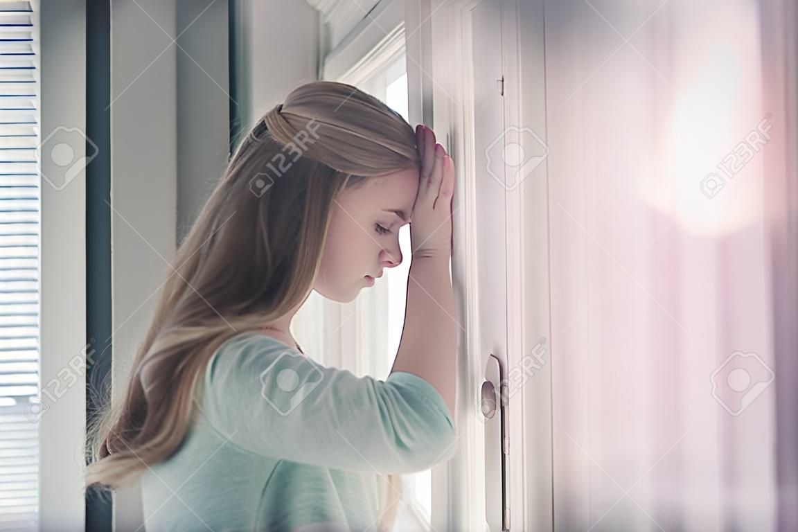 Praying or meditating. Pretty girl or young woman with closed eyes on cute face and blond, long hair standing at window on sunny day.