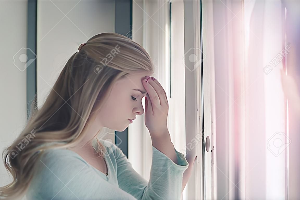 Praying or meditating. Pretty girl or young woman with closed eyes on cute face and blond, long hair standing at window on sunny day.