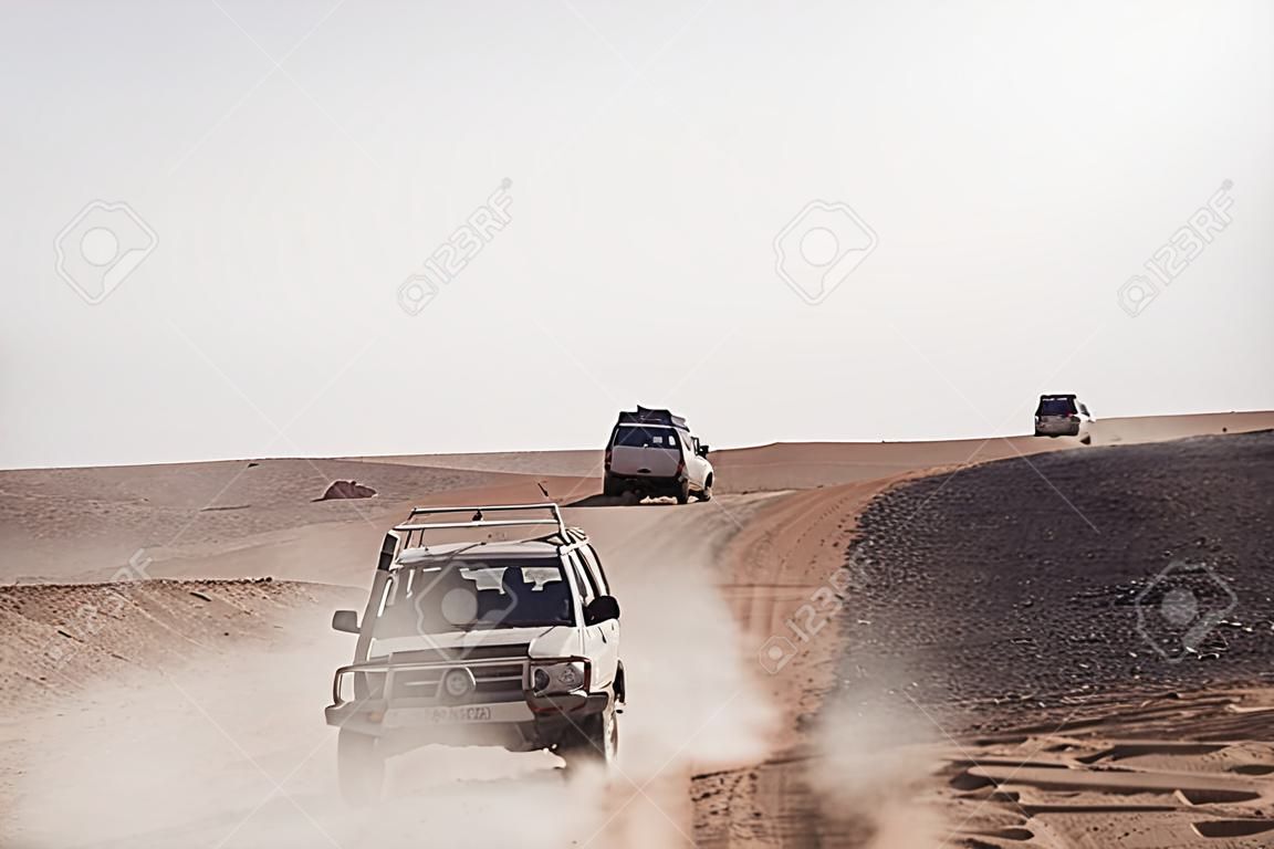 Vehicles bashing through desert hills on dusty sand road on white sky background.