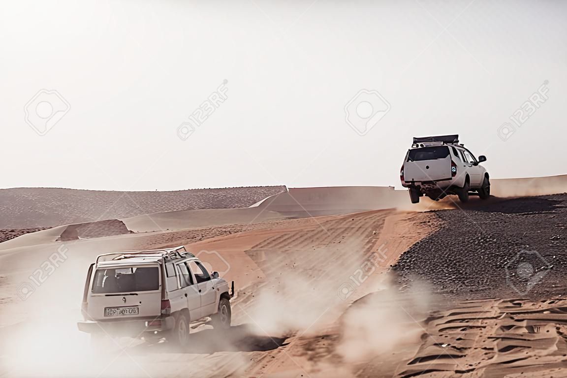 Vehicles bashing through desert hills on dusty sand road on white sky background.