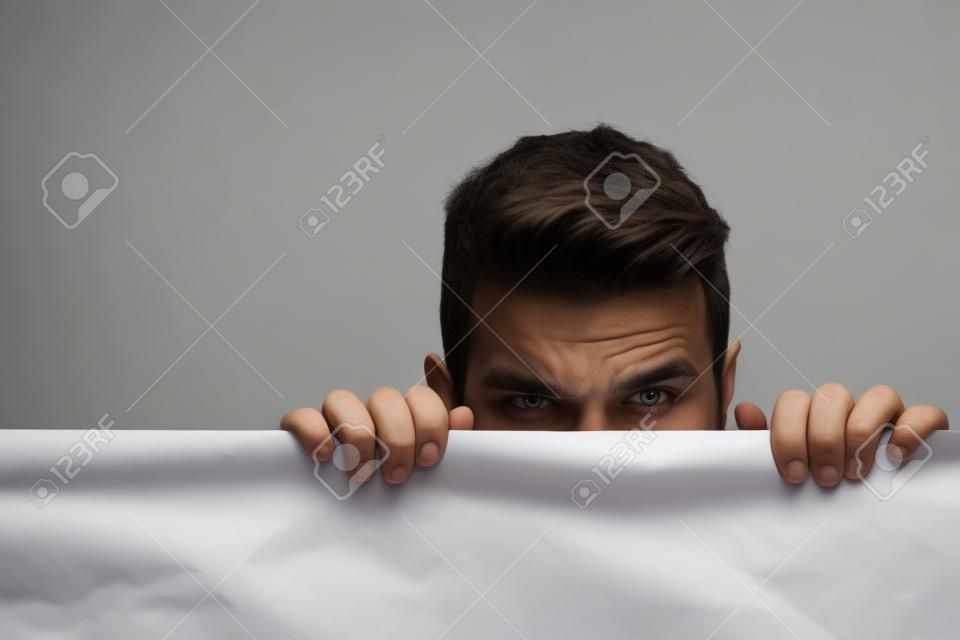 young man with scared eyes on emotional face with long hair behind white paper sheet in studio on grey background, copy space