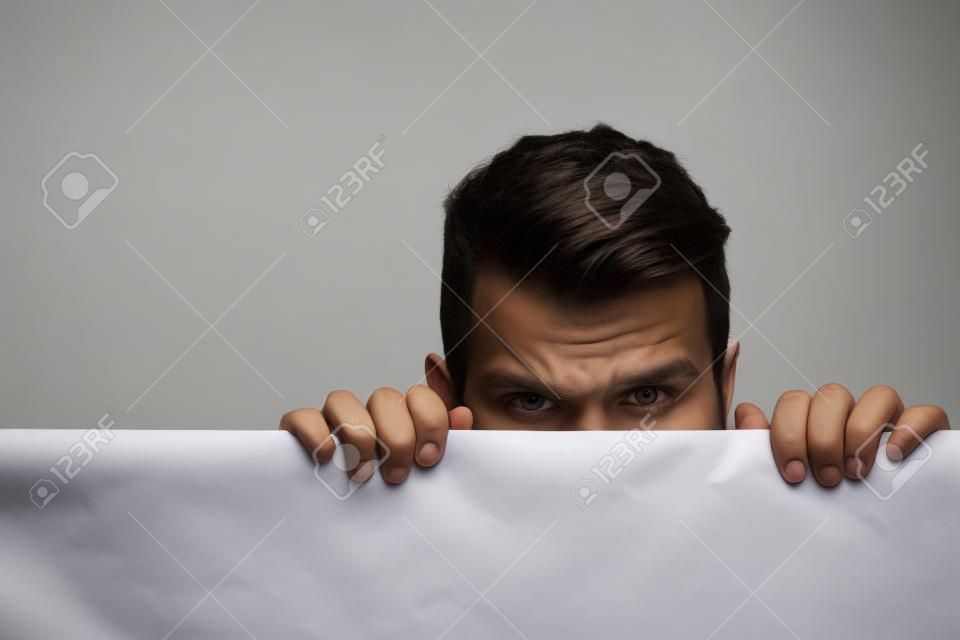 young man with scared eyes on emotional face with long hair behind white paper sheet in studio on grey background, copy space
