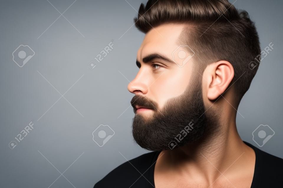 Macho com perfil facial barbudo e elegante pose de cabelo em fundo cinza. Conceito de barbeiro, barbearia, cabeleireiro ou salão de beleza, espaço de cópia