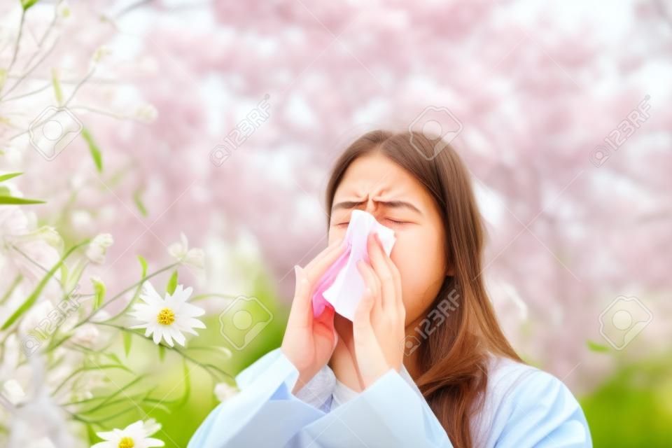Cold flu season, runny nose. Flowering trees in background. Young girl sneezing and holding paper tissue in one hand and flower bouquet in other. Flu. Allergy season.
