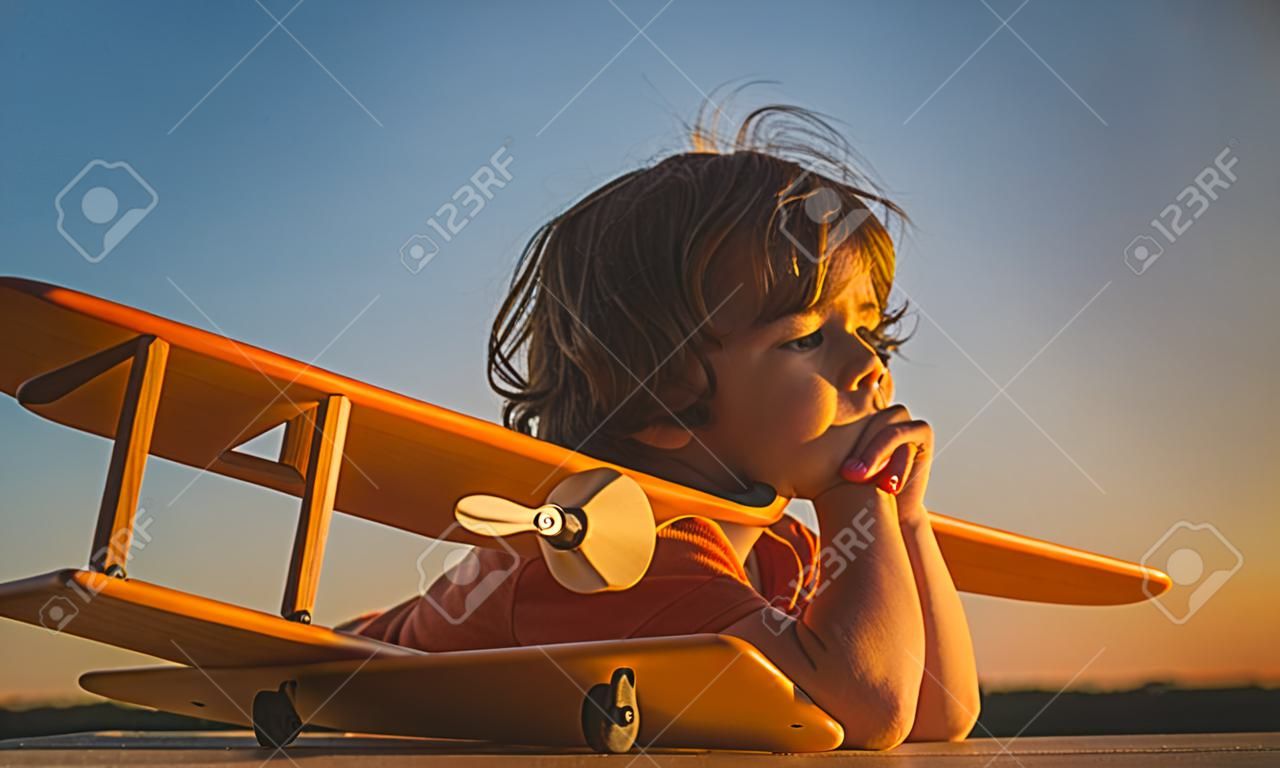 Child dreams and imagination concept. Dreaming child playing with toy wooden airplane against summer sky background. Retro toned in sunset.