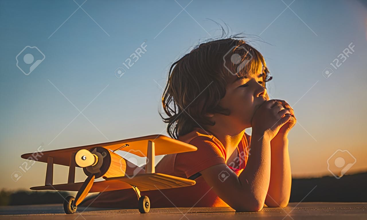 Child dreams and imagination concept. Dreaming child playing with toy wooden airplane against summer sky background. Retro toned in sunset.