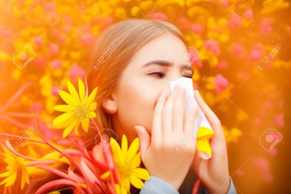 Portrait of an allergic woman surrounded by seasonal flowers wearing bright yellow sweater. Charming girl is blowing her nose near autumn yellow flowers in bloom.