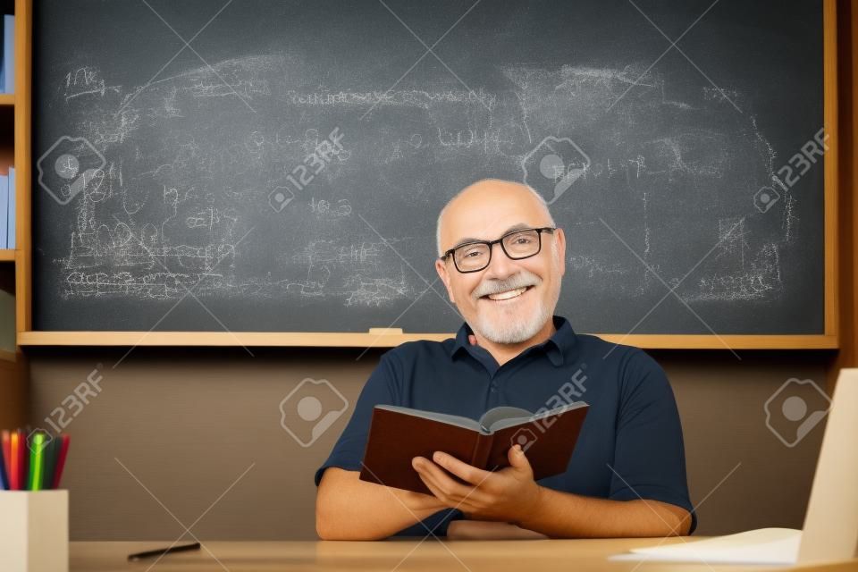 Man mature teacher. Portrait of happy senior tutor in glasses with book on the blackboard in class at high school or collage. Old student study in university.