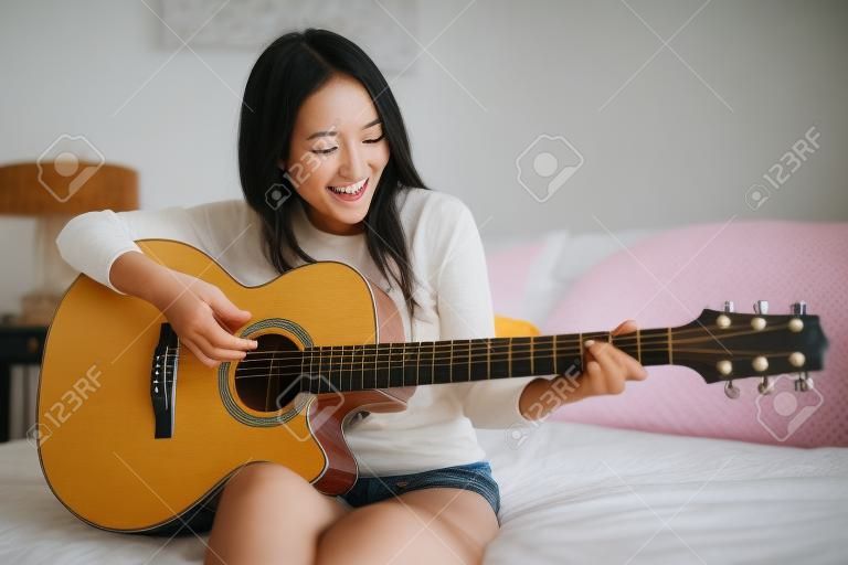 かわいい、幸せな笑みを浮かべてアジアの女の子の寝室でアコースティック ギターを演奏