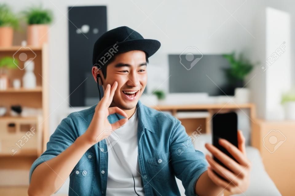 Young happy Asian man talking video call via smartphone wearing headphones at home