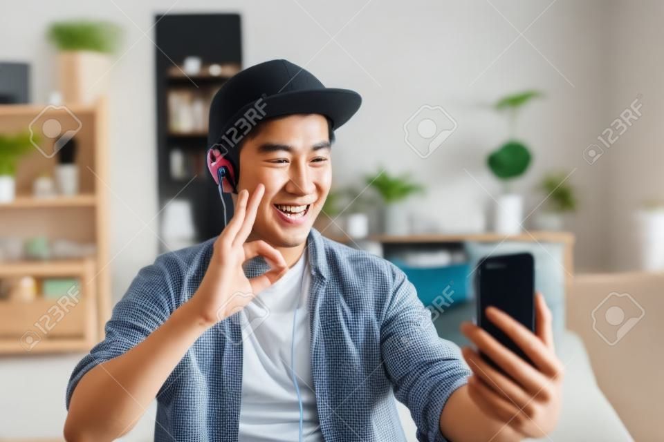 Video chiamata di conversazione del giovane uomo asiatico felice tramite le cuffie d'uso dello smartphone a casa