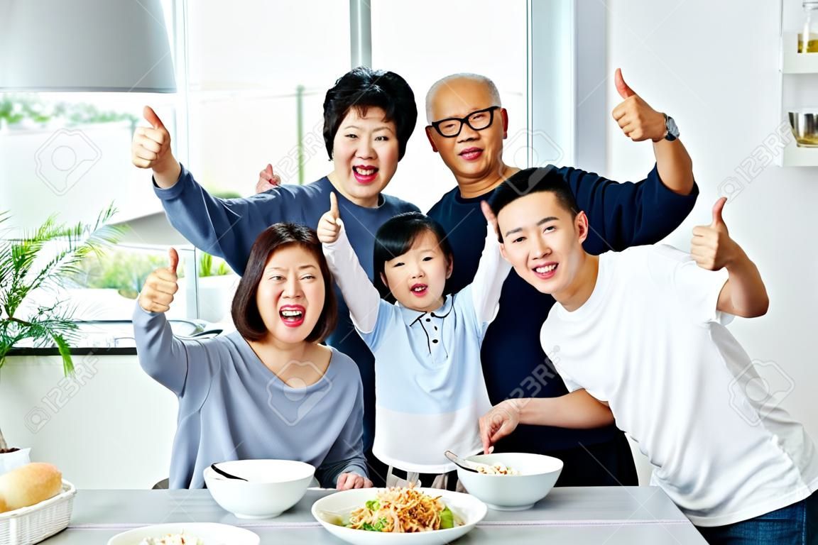 Üç kuşaktan oluşan geniş Asyalı aile birlikte yemek yiyor ve evde mutlulukla baş parmaklarını gösteriyor