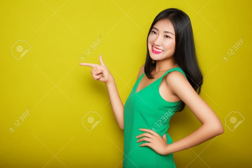 Asiatische niedliche Frau, die Hände zu leerem Kopierraum zeigt und Arme in die Seite gestemmt auf gelbem Hintergrund aufwirft
