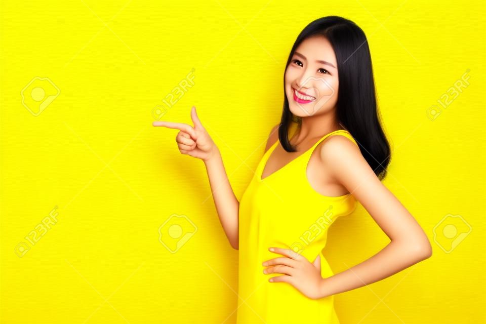 Asiatische niedliche Frau, die Hände zu leerem Kopierraum zeigt und Arme in die Seite gestemmt auf gelbem Hintergrund aufwirft