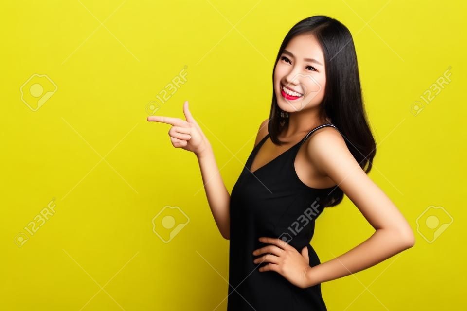 Donna asiatica carina che indica le mani per svuotare lo spazio della copia e posa le braccia sui fianchi isolato su sfondo giallo