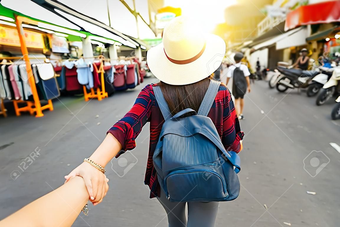 Back side of Young Asian traveling women leading by holding her boyfriend hand in Khaosan Road walking street in evening at Bangkok, Thailand, traveleling together and tourist with follow me concept