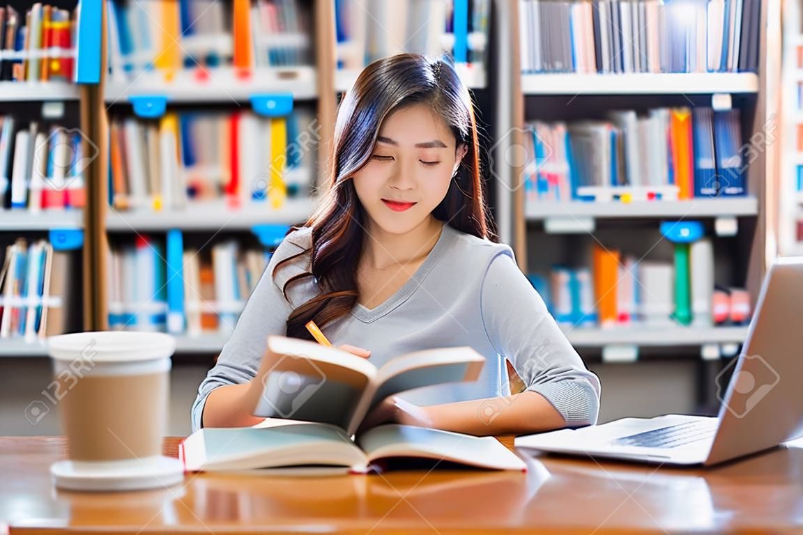 Azjatycki młody student w swobodnym garniturze odrabia pracę domową i korzysta z technologicznego laptopa w bibliotece uniwersytetu lub uczelni z różnymi książkami i urządzeniami stacjonarnymi na tle półki z książkami powrót do szkoły