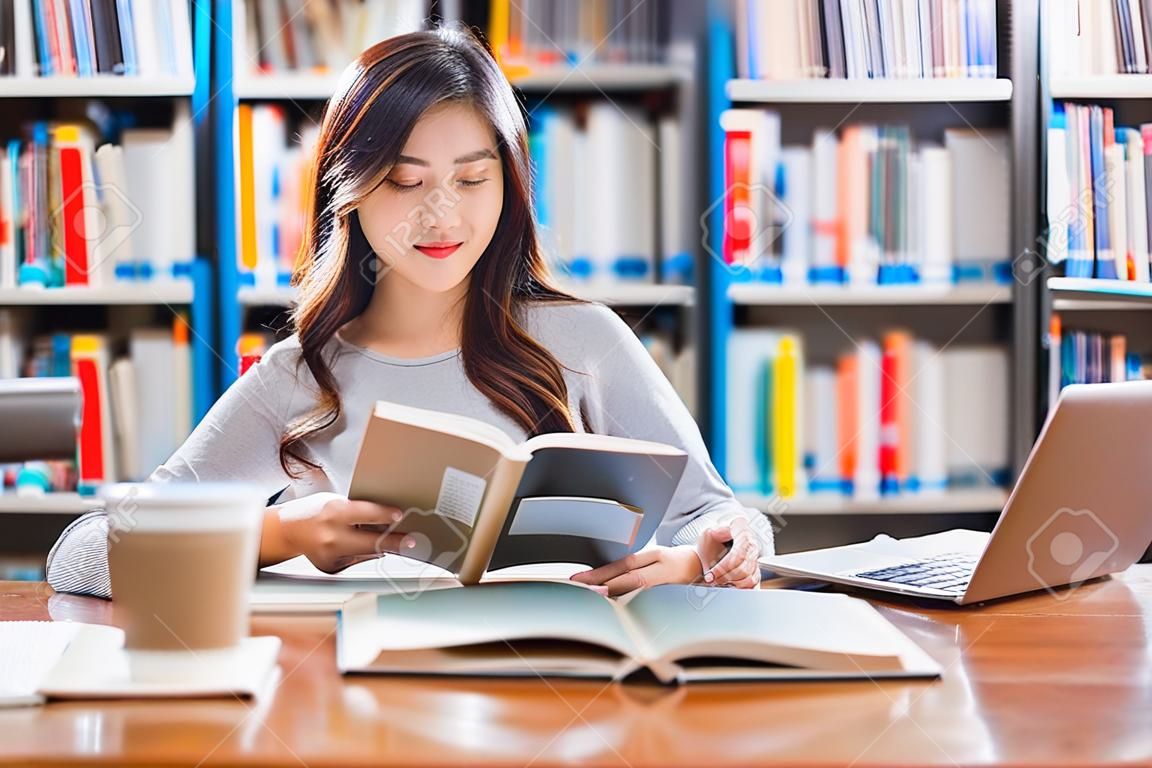 Azjatycki młody student w swobodnym garniturze odrabia pracę domową i korzysta z technologicznego laptopa w bibliotece uniwersytetu lub uczelni z różnymi książkami i urządzeniami stacjonarnymi na tle półki z książkami powrót do szkoły