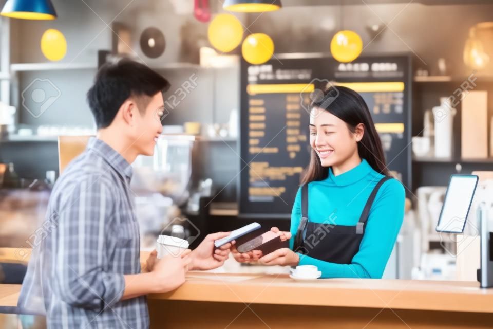 Uomo del cliente asiatico che paga tramite canale contactless tramite applicazione di mobile banking al proprietario di una piccola impresa asiatica al tavolo nella caffetteria, titolare di una piccola impresa e avvio nel concetto di caffetteria