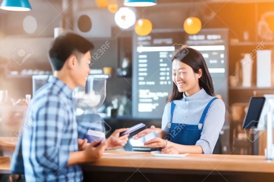 Uomo del cliente asiatico che paga tramite canale contactless tramite applicazione di mobile banking al proprietario di una piccola impresa asiatica al tavolo nella caffetteria, titolare di una piccola impresa e avvio nel concetto di caffetteria