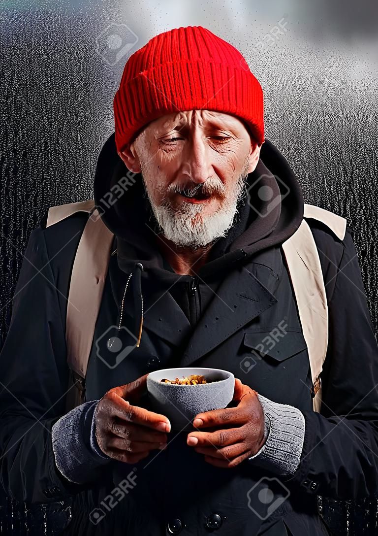 Hombre mendigo llora por falta de comida. Sueña con refugio y comida. Hombre sin hogar de pie bajo la lluvia, mira a la cámara