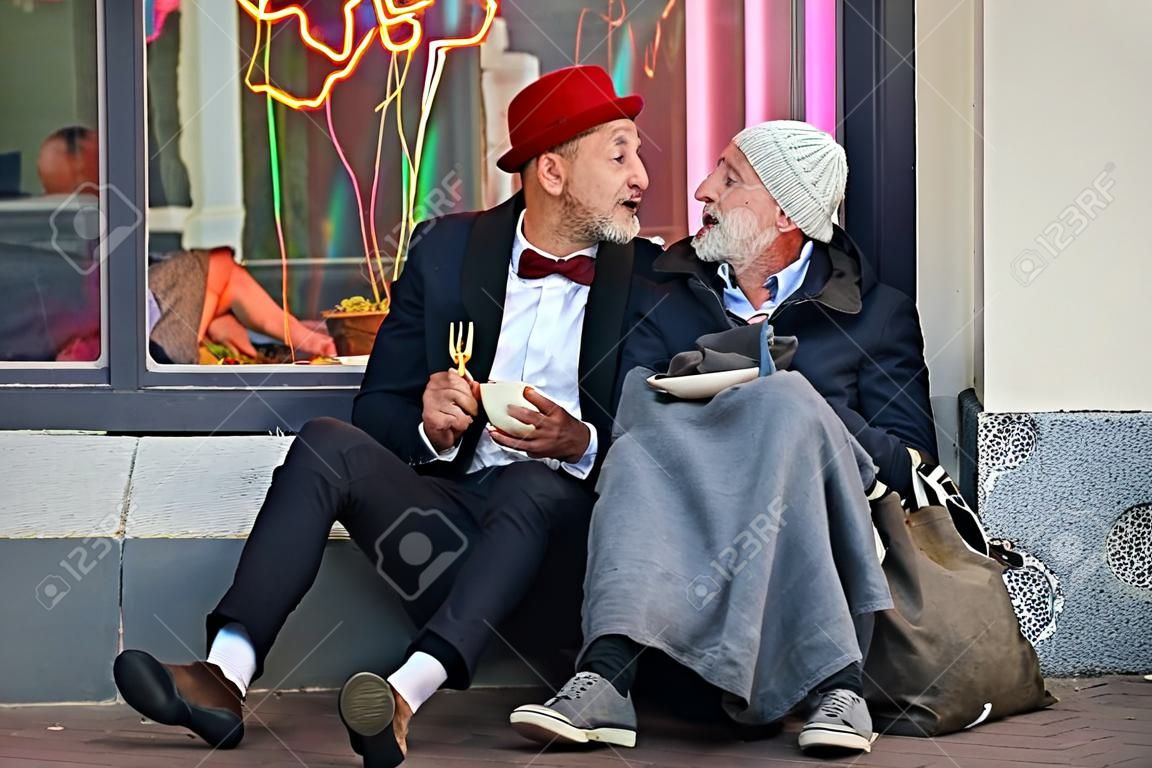 Stijlvolle kantoormedewerker en bedelaar man zitten te eten op straat en spreken. Man zonder smoking, arme man in oude kleren