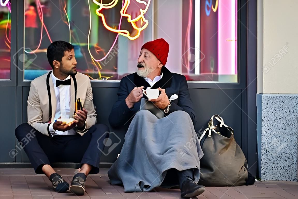 Stijlvolle kantoormedewerker en bedelaar man zitten te eten op straat en spreken. Man zonder smoking, arme man in oude kleren